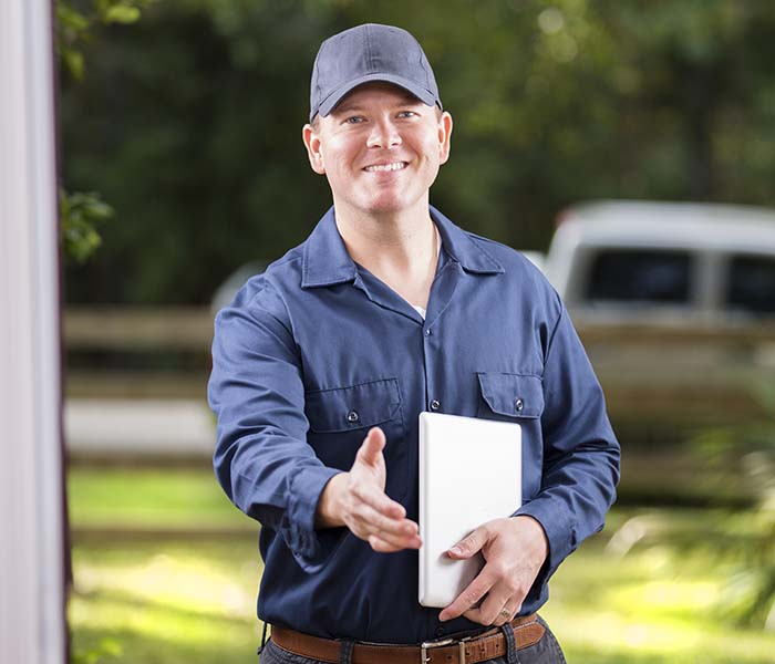Mid-adult repairman or blue collar/service industry worker makes service/house call at customer's front door. He holds his digital tablet with work invoices and reaches out to shake hands. Inspector, exterminator, electrician. He wears a navy colored uniform. Service truck seen in background parked on road.