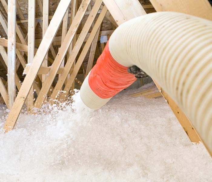 A worker is spraying blown fiberglass insulation between attic trusses at a residential construction site. The completed application will be 22” deep providing an insulation value of R-60.